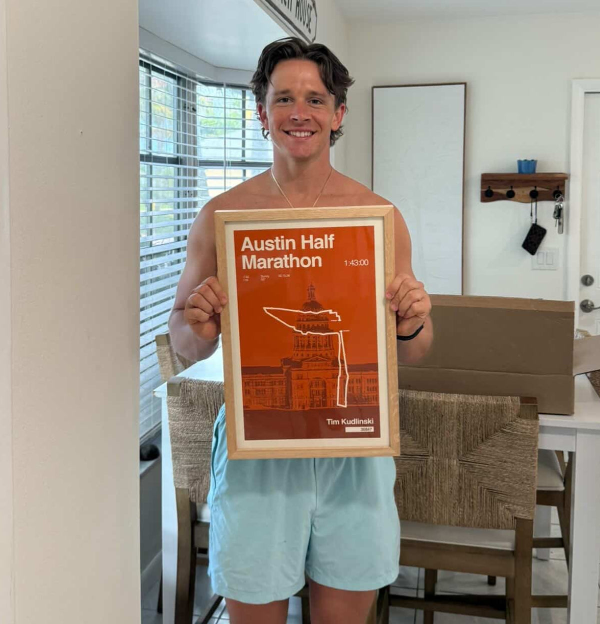 Man holding a framed poster of the Austin Half Marathon in a room with a window and furniture.